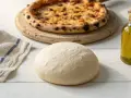 Świeże ciasto na pizzę, gotowe do pieczenia, obok butelki oliwy i upieczonej pizzy.