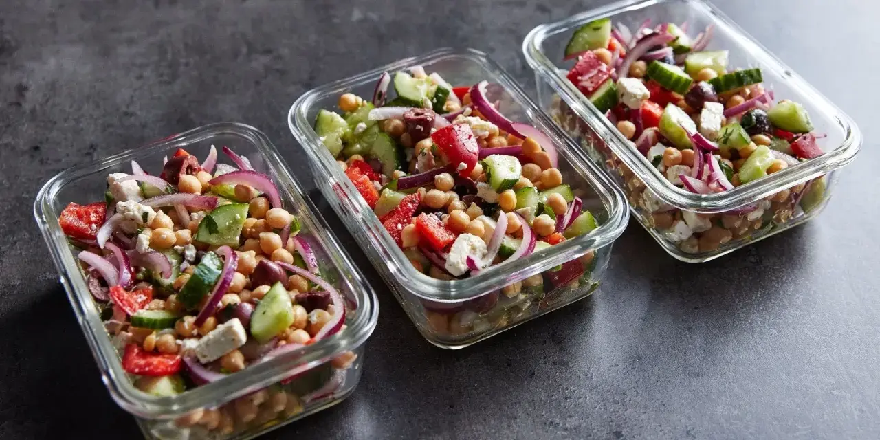 Trzy szklane pojemniki wypełnione kolorową sałatką z ciecierzycą, ogórkiem, pomidorem, czerwoną cebulą i fetą. Idealna na lunch.