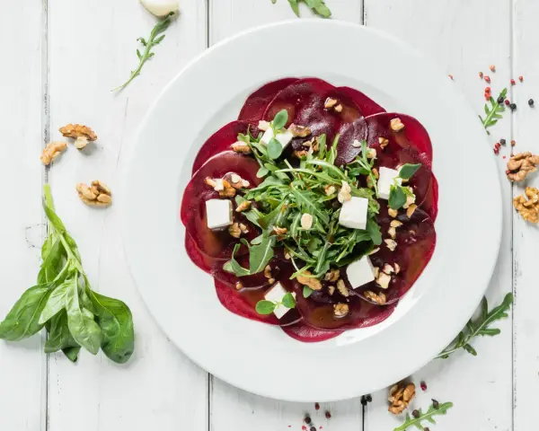 Eleganckie carpaccio z buraka z rukolą, orzechami włoskimi i kostkami sera feta na białym talerzu.