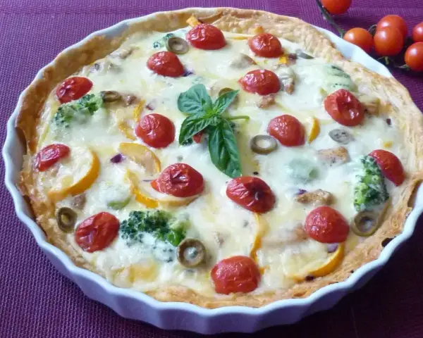 Pyszna tarta na słono z brokułami, pomidorkami koktajlowymi i oliwkami, udekorowana listkiem bazylii.