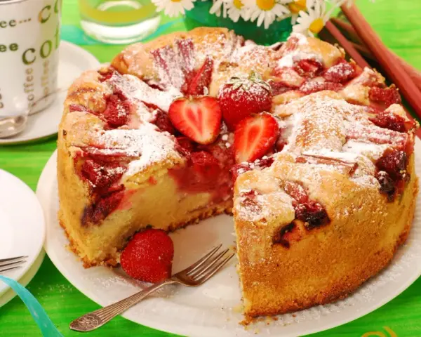 Pyszne ciasto drożdżowe z truskawkami, posypane cukrem pudrem, czeka na spróbowanie. Obok filiżanka kawy i świeże truskawki.