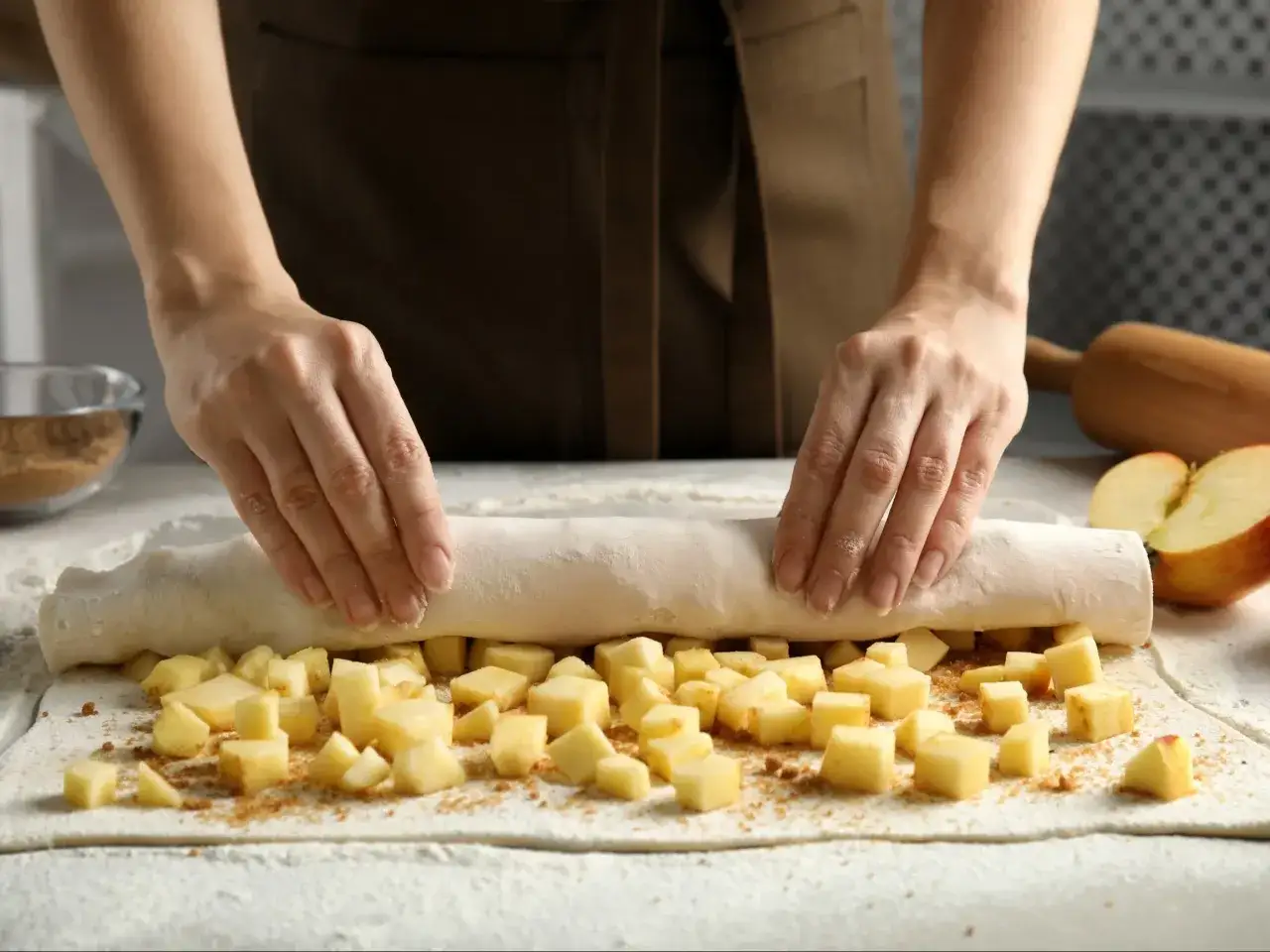 Ręce zwijają ciasto z jabłkami, tworząc pyszny strudel z jabłkami. Obok leżą jabłka i wałek.