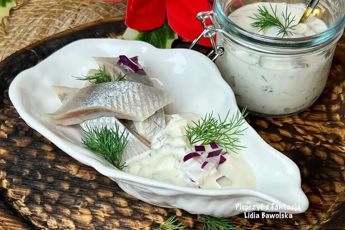 Pyszne śledzie w śmietanie z koperkiem i czerwoną cebulką, podane w białej miseczce. Obok słoiczek z sosem.