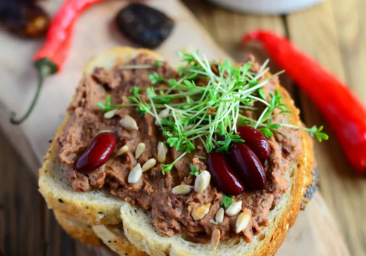 Pyszna pasta z czerwonej fasoli na chlebie, ozdobiona kiełkami i nasionami. Obok leżą czerwone papryczki chili.